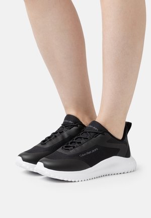Sneakers nere Calvin Klein Jeans con suole bianche, indossate a piedi nudi, viste di lato e di fronte su uno sfondo neutro.