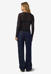 Ženska z blond lasmi, oblečena v črno prozorno majico z dolgimi rokavi, temno modre široke kavbojke in bele superge, obrnjena stran na belem ozadju.