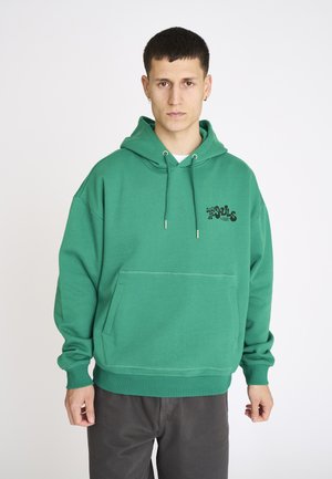 Ung mand iført en grøn hættetrøje med frontlomme og sort tekst på brystet, stående mod en ensfarvet hvid baggrund.