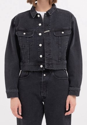 Femme portant une veste en denim noir courte avec des boutons argentés et un pantalon en denim noir assorti à taille haute, avec une chemise blanche en dessous.