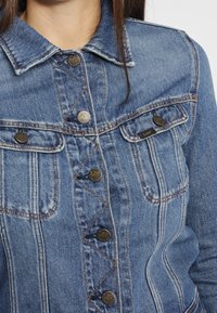 Chaqueta de mezclilla en un azul claro con cierre de botones en la parte delantera, bolsillos en el pecho y detalles de costura visibles. Diseño de collar clásico y ajuste regular.