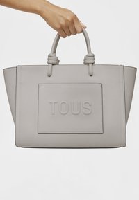 Sac à main en cuir texturé gris avec poignées doubles, arborant un logo "TOUS" en relief sur un panneau rectangulaire.
