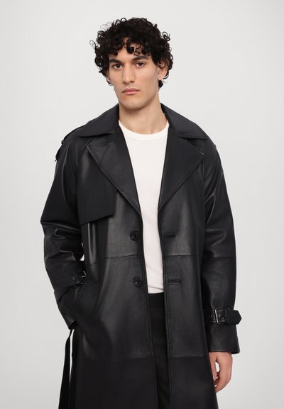 Svart läder trench coat med breda slag, dubbelknapprad och justerbara spännen på ärmarna. Slät textur och skräddarsydd passform.