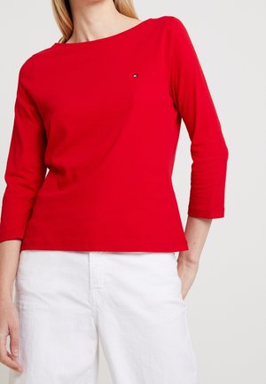 Femme portant un haut à manches longues rouge avec un petit logo sur la poitrine et un pantalon blanc, debout avec une main dans la poche.