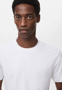 Camiseta blanca de algodón con mangas cortas y cuello redondo. Textura suave, diseño ajustado. Tejido liso, sin patrones ni acentos visibles.