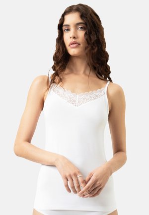 Frau mit langen, lockigen Haaren, die ein weißes Camisole mit Spitzenbesatz trägt, vor einem schlichten, hellen Hintergrund stehend, die Hände sanft ineinandergelegt.