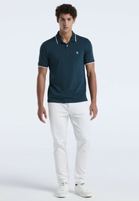 Dunkelblaues Poloshirt mit weißen Akzenten, kurzen Ärmeln und kleinem Logo. Kombiniert mit weißen Hosen und weißen Sneakers.