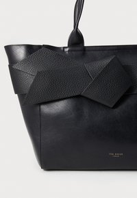Sac cabas en cuir noir avec un accent texturé géométrique et une finition lisse. Inclut un logo doré en bas.