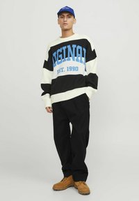Maglione a righe nere e crema con testo blu "Original EST. 1990." Abbinato a pantaloni neri e stivali color cammello con lacci.