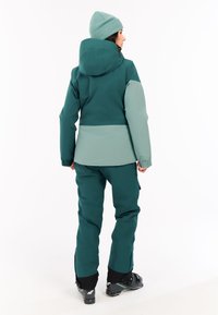 Zweiteiliges Outdoor-Outfit bestehend aus einer Kapuzenjacke und einer Hose in einem gedämpften Grünton. Die Jacke hat kontrastierende hellgrüne Bereiche.