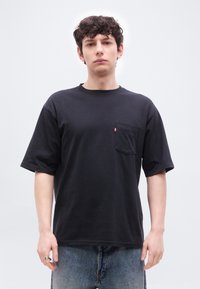 Νεαρός άνδρας με κατσαρά μαλλιά, φοράει μαύρο κοντομάνικο μπλουζάκι με μπροστινή τσέπη και μπλε τζιν, στέκεται μπροστά σε απλό λευκό φόντο.