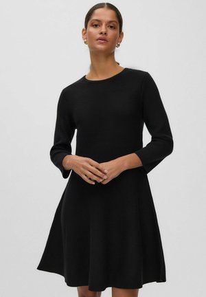 Femme portant une robe noire à manches longues, longueur genou, avec un col rond, debout, les mains jointes devant un fond uni.