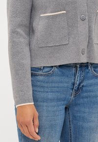 Γκρι πλεκτό cardigan με δύο μεγάλες τσέπες, ανοιχτό κρεμ χρώματα και κουμπωτό μπροστινό μέρος. Συνδυασμένο με μπλε τζιν, αναδεικνύοντας την υφή.