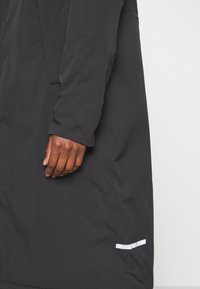 Cappotto nero con maniche lunghe, texture liscia, leggero lucido, con una striscia di accento bianca vicino all'orlo e un design complessivo minimale.
