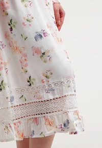 Vestido de gasa floral con base blanca, que presenta patrones en rosa, púrpura y verde. Incluye detalles de encaje y un dobladillo con volantes en la parte inferior.