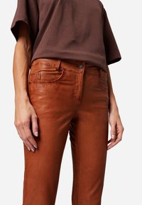 Bruine leren broek met een aansluitend ontwerp, voorzakken en ton-sur-ton stiksels. Soepele textuur met een lichte glans en een knoopsluiting.