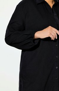 Maglietta nera con chiusura frontale a bottoni, texture rigata e maniche arricciate. Il tessuto appare morbido e leggermente lucido.