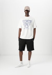 Balta medvilninė marškinė su mėlynu Niujorko "Yankees" grafiniu estampu, juodos tinklinės šortai ir baltos sportinės batu. Šviesiai ruda kepurė puikiai užbaigia laisvalaikio aprangą.