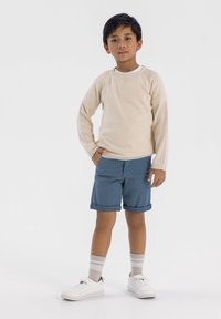 Ljusbeige stickad tröja, blå rullade shorts, vita sneakers och ljusrandiga strumpor. Kille står självsäkert med ena handen i fickan.