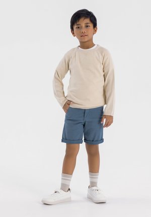 Hellbeige gestrickter Pullover, blaue Shorts mit Umschlag, weiße Sneakers und hell gestreifte Socken. Junge steht selbstbewusst mit einer Hand in der Tasche.