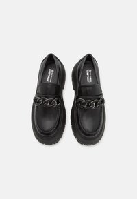 Mocassins en cuir noir avec une semelle épaisse, dotés d'un accent en chaîne en argent proéminent. Texture lisse et design à bout rond.