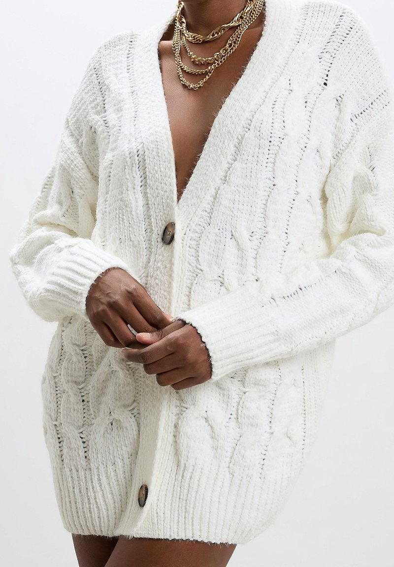 Personne à la peau foncée portant un cardigan blanc épais en maille torsadée avec de gros boutons et des colliers à chaînes dorées superposées.