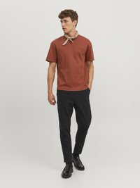 Rostfarbiges Baumwoll-T-Shirt mit kurzen Ärmeln und einem kleinen schwarzen Druck, kombiniert mit schwarzen Slim-Fit-Hosen und schwarzen Slippern. Heller Schal um den Hals gelegt.