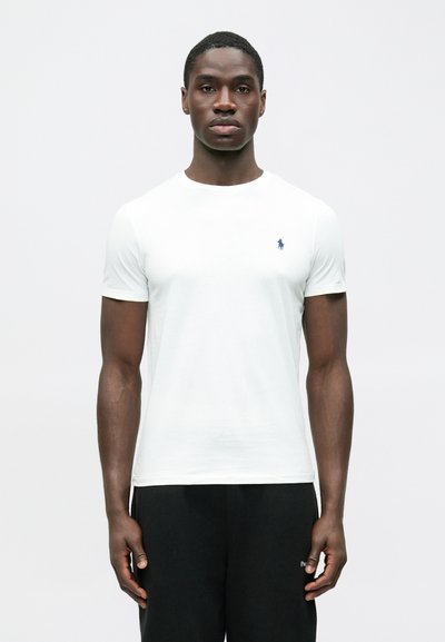 T-shirt branca de manga curta feita de algodão, com um pequeno logótipo azul-marinho no lado esquerdo do peito, combinada com calças pretas. Design simples, cores sólidas.