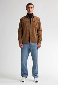 Chemise marron à boutons avec deux poches poitrine, portée par-dessus un col roulé noir, associée à un jean bleu clair et des baskets blanches.