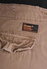 Bermudas de carga beige con un bolsillo acolchado, detalles cosidos y una etiqueta negra con texto naranja; hechas de un tejido duradero.