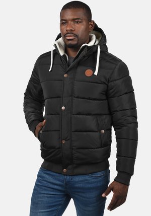 Black puffer jacket with horizontal quilted design, front buttons, drawstring hood, and fleece-lined collar. Brown leather logo patch on chest.