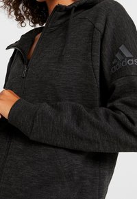 Felpa nera con zip realizzata in tessuto testurizzato, dotata di cappuccio regolabile e di un discreto logo Adidas sulla manica sinistra.