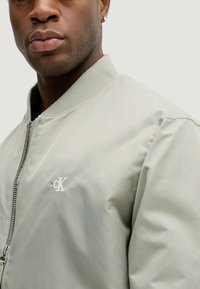 Giacca bomber grigio chiaro con chiusura a zip, colletto a costine e logo Calvin Klein sottile. Presenta una texture liscia e leggermente lucida.