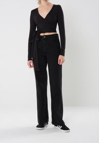 Frau in einem schwarzen langärmeligen Wickel-Crop-Top, hoch taillierten schwarzen Jeans und schwarzen Sneakers mit weißen Streifen, die vor einem schlichten weißen Hintergrund steht.