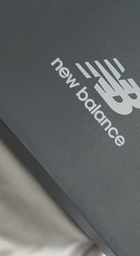 Μαύρο κουτί παπουτσιών με υφή στην επιφάνεια, με λευκό λογότυπο "new balance" και στυλιζαρισμένο σχέδιο "N" στην κορυφή.