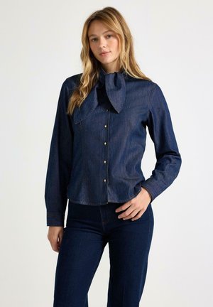 Donna che indossa una camicia di jeans blu scuro con collo a fiocco grande e pantaloni di jeans blu scuro abbinati, in piedi su sfondo bianco.