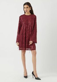 Viininpunainen paljettimekko, jossa pitkät hihat ja hapsut, pyöreä pääntie ja väljä istuvuus, yhdistetty mustiin stilettokorkoihin.