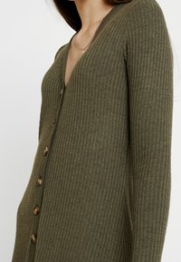 Cardigan côtelé vert olive avec encolure en V, doté de six boutons en bois sur le devant et de manches longues. Texture douce et silhouette ajustée.