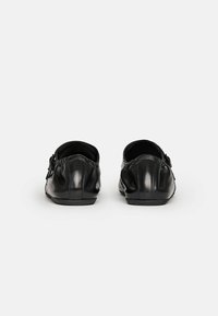 Scarpe slip-on in pelle nera con una superficie liscia, punta arrotondata e un dettaglio arricciato unico sul retro. Hardware di accento sottile sui lati.