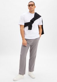 Witte T-shirt met korte mouwen, grijze op maat gemaakte broek, zwarte sweatshirt over de schouder gedrapeerd, heldere bril, witte sneakers.
