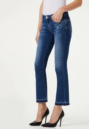 Kobieta w niebieskich, krótkich obcisłych dżinsach z postrzępionymi końcówkami i czarnych szpilkach, stojąca z jedną ręką w kieszeni.