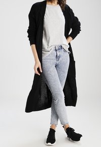 Cardigan lungo nero a maglia indossato sopra una maglietta grigia, abbinato a jeans skinny blu chiaro e sneakers nere. Presenta un fit rilassato e un design aperto.