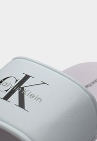 Κοντινό πλάνο ενός λευκού σανδαλιού Calvin Klein με μαύρο λογότυπο "CK" στο λουρί και ανοιχτό μοβ σόλα στο φόντο.