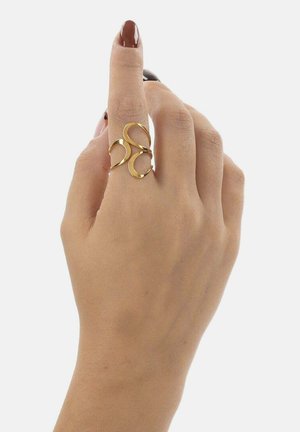 Hand mit dunkelbraun lackierten Nägeln, die einen offenen Goldring mit abstrakten, geschwungenen Formen am Mittelfinger trägt.