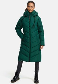 Cappotto imbottito verde, trapuntato, con cappuccio, caratterizzato da cuciture diagonali, chiusura zip e tasche laterali. Indossato con stivali da combattimento neri.