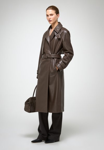Bruine leren trenchcoat met een ceintuur in de taille, brede kraag en zijzakken, gecombineerd met donker op maat gemaakte broeken en spitse zwarte schoenen.