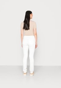 Top de manga corta beige combinado con jeans ajustados blancos. La modelo está de pie frente a un fondo neutro, mostrando las líneas y colores limpios del conjunto.