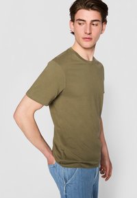 Olivenfarget t-skjorte med korte ermer laget av myk bomull, med rund hals, jevn tekstur og en tettsittende design. Brukt sammen med blå denimjeans.