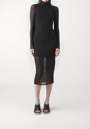 Žena má na sobě přiléhavé černé midi šaty s průsvitnými dlouhými rukávy a průsvitnou spodní částí sukně, doplněné černými otevřenými nazouvacími sandály.