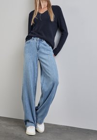 Jean large bleu clair à taille haute, avec un design classique à cinq poches, associé  à un pull en V bleu marine foncé et des baskets blanches.